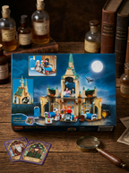 A boxed Harry Potter collectible wizard castle set displayed on a wooden table with vintage books and glass bottles in the background. The box shows a detailed castle scene at night with a full moon, and small accessory cards and a magnifying glass are placed in front.